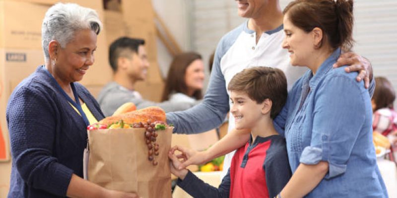 Multi-ethnic, mixed age group of volunteers work together at food bank.  They pack sacks and boxes of food for needy people in their community.  Senior woman gives sack full of groceries to needy family.